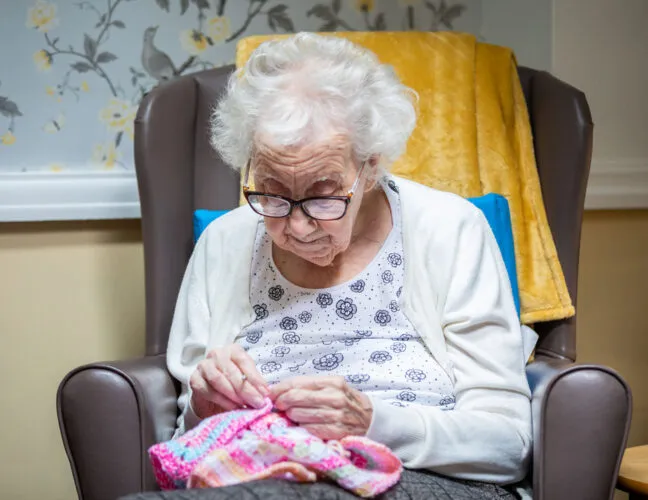 An elderly resident knitting in a comfy chair at St Oswalds House care home - Maven Healthcare